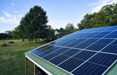 Solar panels on a roof in the foreground, with a house, large trees, and a grassy yard under a partly cloudy sky in the background.