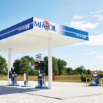 A small outdoor gas station with a white canopy displaying MFA Oil branding offers seamless fuel experiences. Featuring two fuel pumps surrounded by a concrete area, the site boasts recent payment upgrades amid trees and a clear blue sky with scattered clouds in the background.