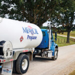 A blue and white propane delivery truck labeled MFA Oil, driven by a professional with Federal Entry-Level Driver Training (ELDT), travels down a rural gravel road surrounded by grassy fields and large trees.
