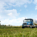 A blue propane delivery truck drives on a grassy field under a partly cloudy sky. The vehicle is prominently displayed in the foreground, with greenery and a fence visible in the background.