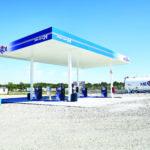 A bright, sunny day at a modern gas station featuring multiple fuel pumps under a white and blue canopy, with visible signage for Marathon. In the background, a large fuel storage tank is seen against a clear blue sky.