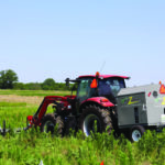 A red tractor is towing a large agricultural machine in a green field under a clear blue sky. The machine has various equipment attached and is surrounded by tall plants. Trees are visible in the distant background.