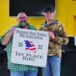 Two men proudly hold a sign reading Missouri High School BBQ Association 2022 1st Place Ribs at an Osceola event. The man on the left sports a green plaid shirt, while the man on the right dons a camo jacket. Behind them, a yellow wall and counter set the scene.