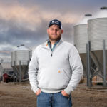 A person in a gray sweater and blue jeans stands confidently in front of large metal silos on a farm. The sky is cloudy, and the ground is gravelly, indicating a rural setting.