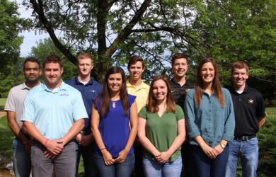 A group of nine interns stands outdoors, smiling at the camera. Casually dressed, they are positioned in two rows with trees and greenery in the background.