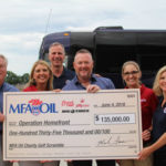 A group of six people is happily holding a large check for $135,000, made out to Operation Homefront from the MFA Oil Charity Golf Scramble, dated June 4, 2018. A tour bus is visible in the background.