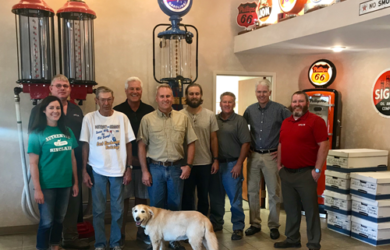 A group of nine people, including a woman, stand indoors in front of vintage gas pumps and signs. A golden retriever is at the front. The room has a retro decor with various colorful signs on the walls.