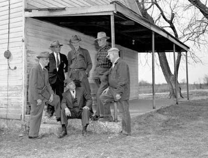 A group of six men in hats and suits gather outside a wooden building. Some stand, while others are seated on the steps. The setting is outdoors, with trees and open land visible in the background. The image is black and white.