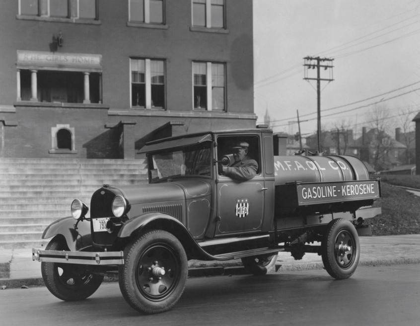 Vintage black and white image of an early 20th-century gasoline and kerosene delivery truck parked on a street. The driver is visible in the truck, and a brick building is in the background with stairs leading up to it.