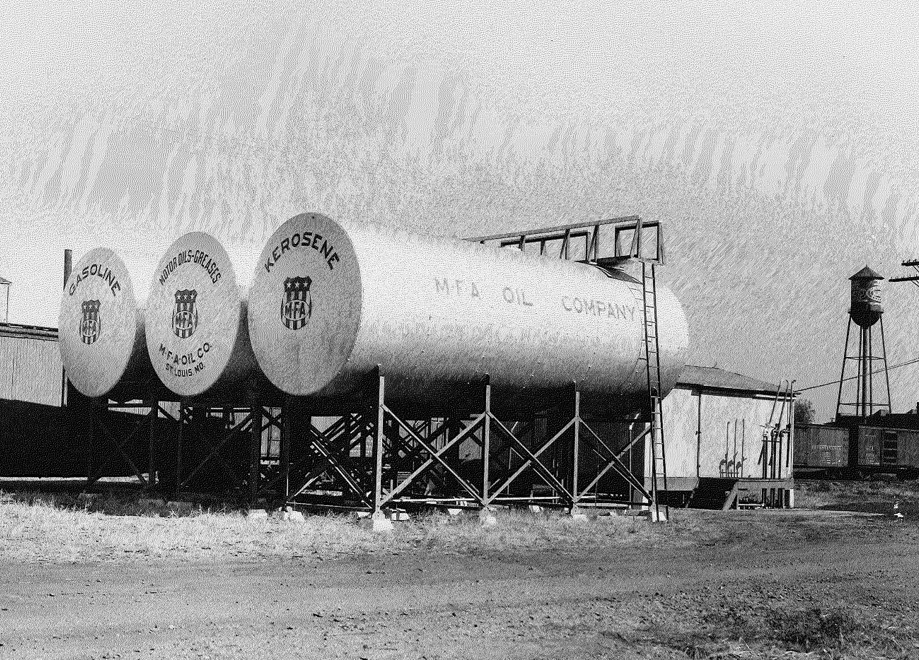 Black and white photo of three large storage tanks labeled Gasoline, Distillate, and Kerosene, belonging to MTA Oil Company. They are elevated on metal stands, with a water tower visible in the background.