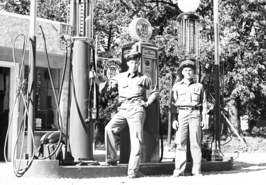 Two men in uniform stand near vintage gas pumps at a station. They appear to be wearing work uniforms, likely indicating they work at the station. The setting looks like its from the mid-20th century, with trees in the background.
