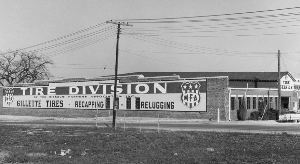 Black and white photo of a building labeled Tire Division of the Missouri Farmers Association Inc. Large signs advertise Gillette Tires, Recapping, and Relugging. Power lines and a tree are visible in the background.