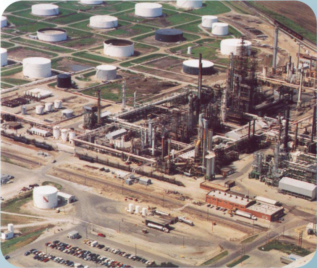 Aerial view of an industrial oil refinery with various tanks and pipelines. Numerous circular storage tanks are spread across the facility. Roads and parked vehicles are visible in the foreground.