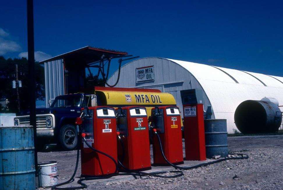 A rural gas station with three red pumps labeled MFA Oil. A blue pickup truck is parked next to a yellow fuel tank. A white, semi-cylindrical building is in the background under a clear blue sky.