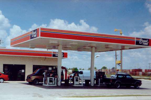 A gas station with a red and white canopy, labeled Break Time, featuring several fuel pumps. A black truck and a white van are parked under the canopy. The building is brick, and the sky is clear with scattered clouds.