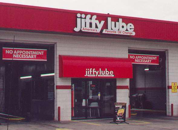 Exterior of a Jiffy Lube service center with red signage. The building displays No Appointment Necessary signs above open service bays. A sandwich board stands outside near the entrance.