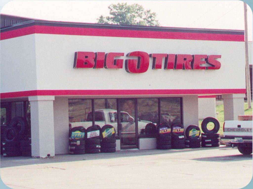 Exterior of a Big O Tires store with a red and white facade. Several tires are displayed outside the window. A Chevrolet truck is parked to the right, and a white pickup truck is reflected in the window.