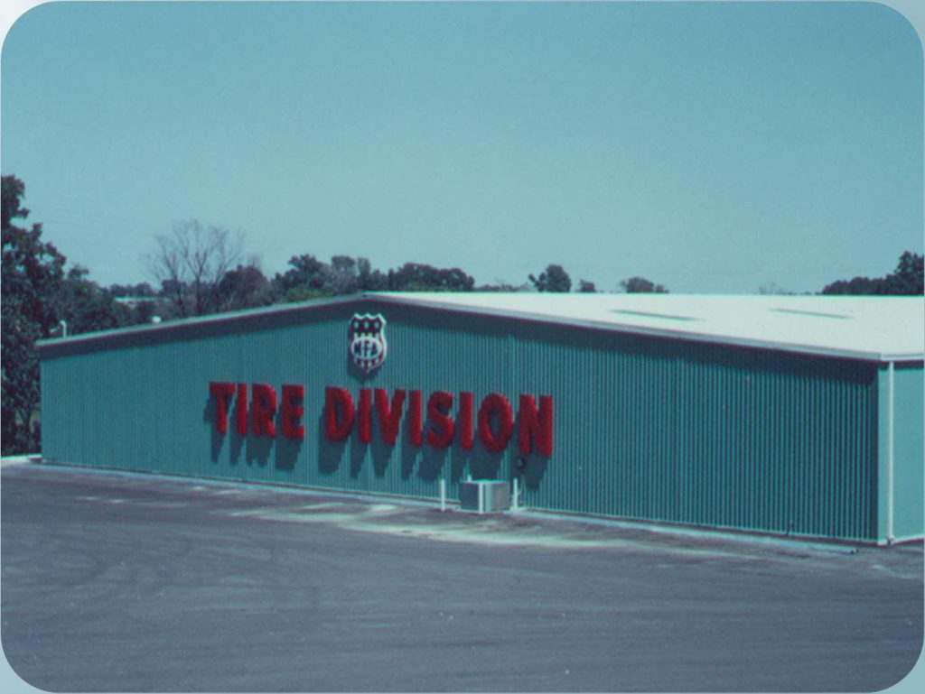 A large warehouse with a teal exterior features bold red letters on the side that read TIRE DIVISION. The building is surrounded by an empty paved area with trees in the background.