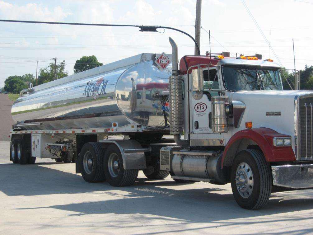 A shiny, silver fuel tanker truck with red accents is parked on a concrete surface. The truck is labeled ETS and features a prominently displayed hazard label. Power lines and trees are visible in the background.