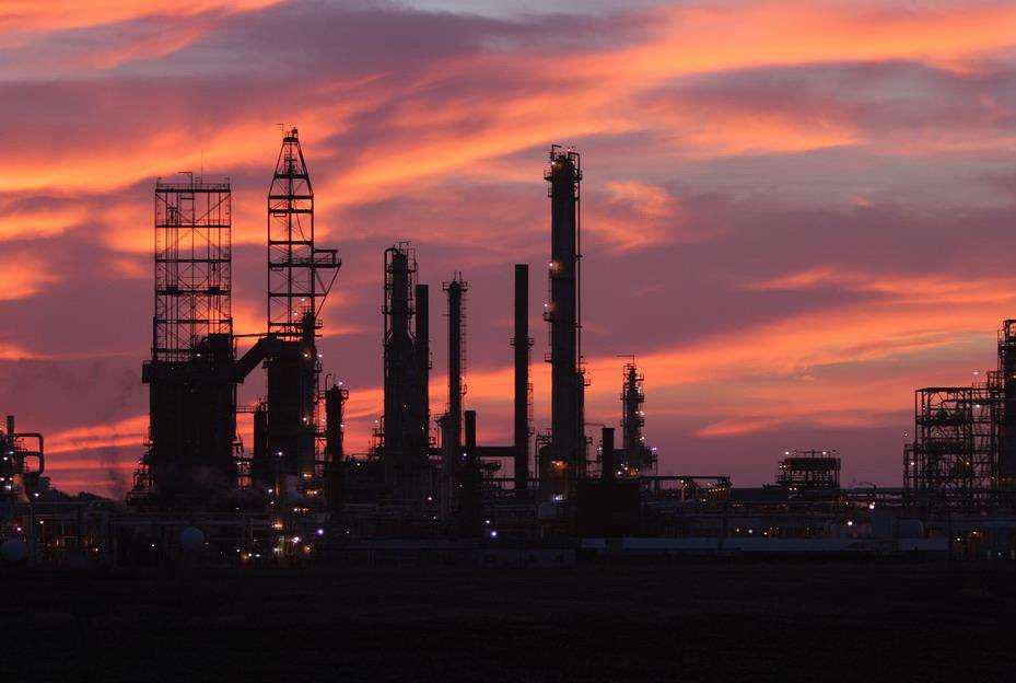 Industrial refinery silhouetted against a vibrant orange and pink sunset sky. Tall chimneys and metal structures are prominent, with the intricate detailing highlighted by the fading light.