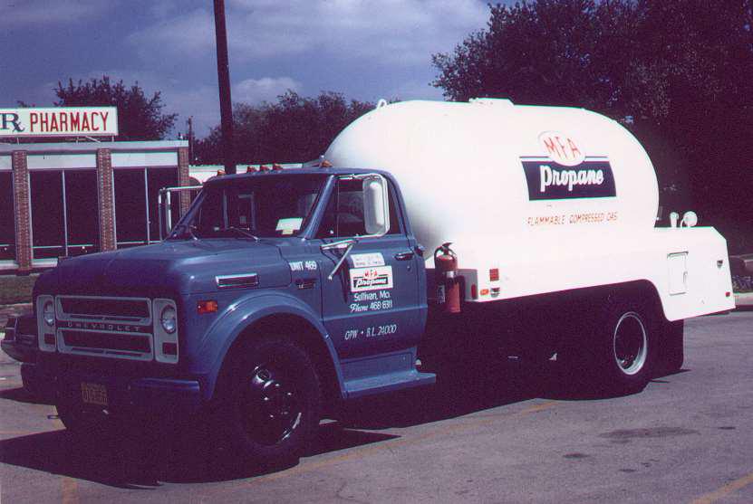 A blue and white propane delivery truck parked near a pharmacy. The truck has a large white tank labeled Propane with a company logo. Trees and part of the pharmacy building are visible in the background.