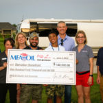 A group of people smiling and holding a large check for $140,000 made out to Operation Homefront. They stand outdoors in front of an RV, dressed casually. The check is from MFA Oil, dated June 3, 2019.