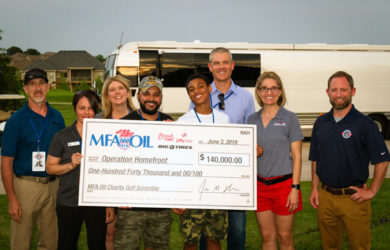 A group of people smiling and holding a large check for $140,000 made out to Operation Homefront. They stand outdoors in front of an RV, dressed casually. The check is from MFA Oil, dated June 3, 2019.
