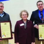 Three people stand indoors. Two men hold framed certificates with gold seals and red, white, and blue ribbons. A woman stands between them, wearing a dark blazer and a white blouse. They appear to be at an official event.