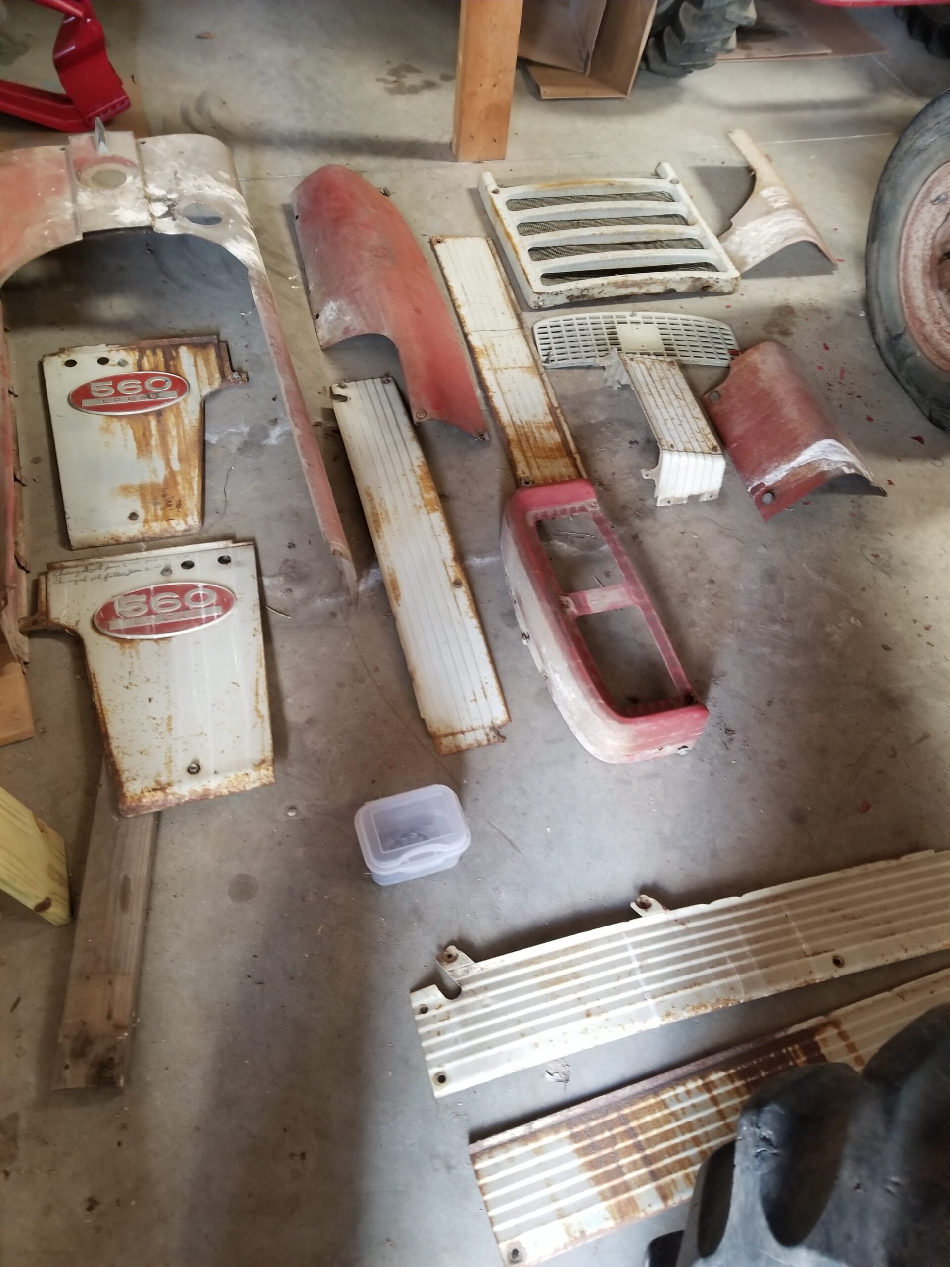 Various rusted and painted metal tractor parts, including grills, fenders, and panels, are spread out on a concrete floor in a workshop.