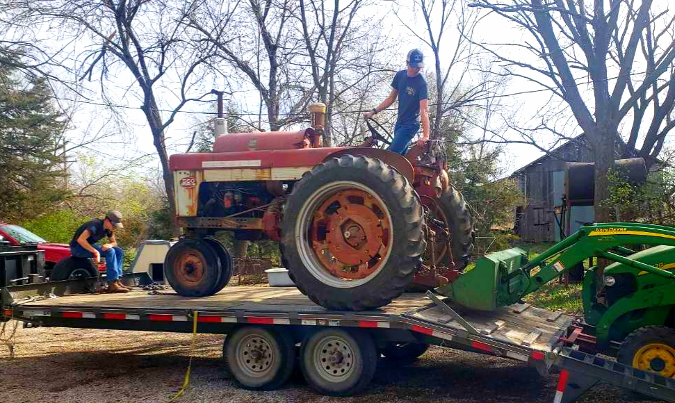 Ty La Row work to load a vintage red tractor onto a tilted trailer using a green loader on a sunny day, with trees and a barn in the background.