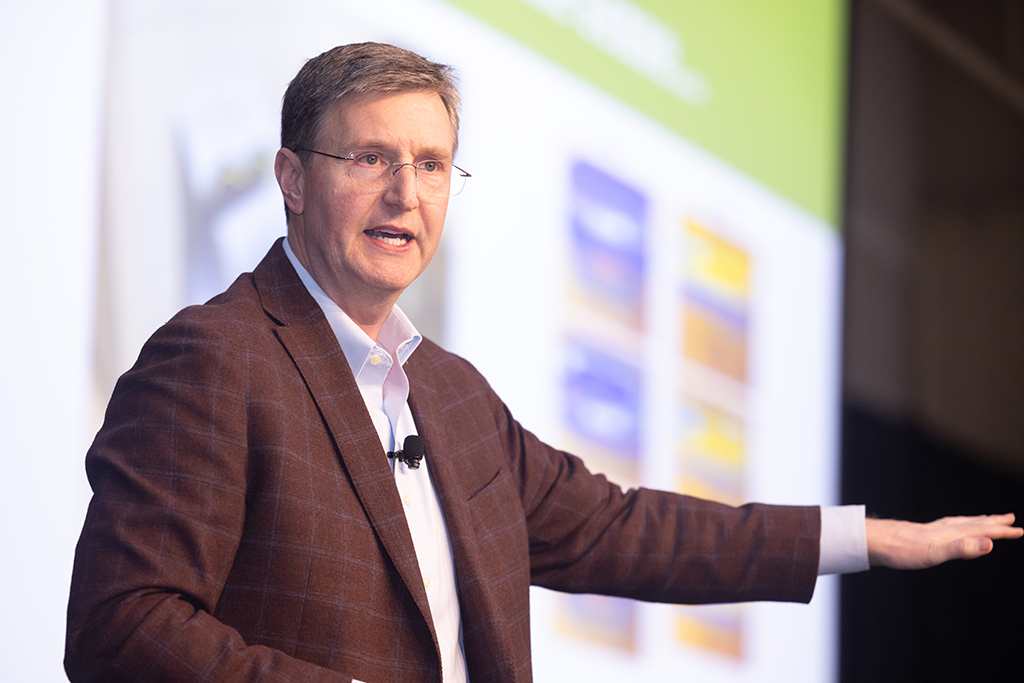 Farmers Versus Foodies author Ray Starling is speaking on stage, gesturing with one hand. Behind him is a blurred presentation screen with colorful graphics.