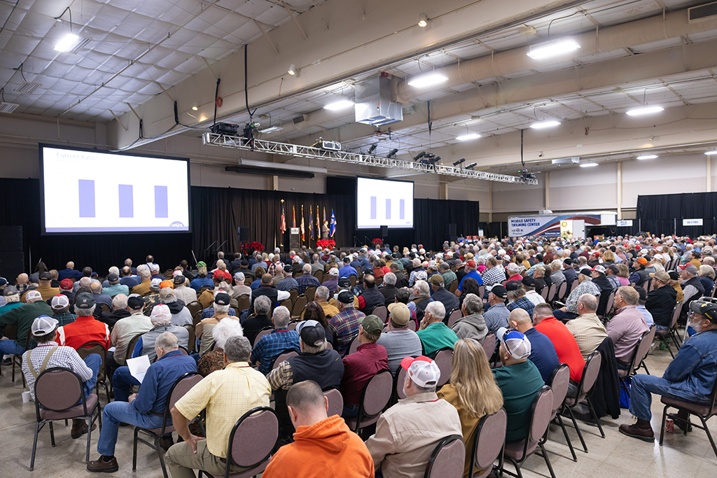 A large group of people seated in rows inside a spacious hall, attending a presentation. Two large screens display a bar graph. Flags and potted plants are on stage. The audience consists mostly of adults, some wearing caps.