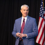 MFA Oil President & CEO, in a blue plaid suit and red tie, speaks on stage, holding a clicker. An American flag is visible in the background. He appears engaged in a presentation or speech.