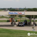 A vehicle is parked at a gas pump under a canopy labeled Platte-Clay Fuels with a grassy landscape and houses in the background. A person is standing next to the vehicle. The Platte-Clay Electric Cooperative logo is displayed in the corner.