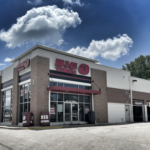 A large building with a Big O Tires sign, featuring a glass entrance, several service bays on the right, and a clear sky with scattered clouds. The exterior is a mix of brick and white paneling.