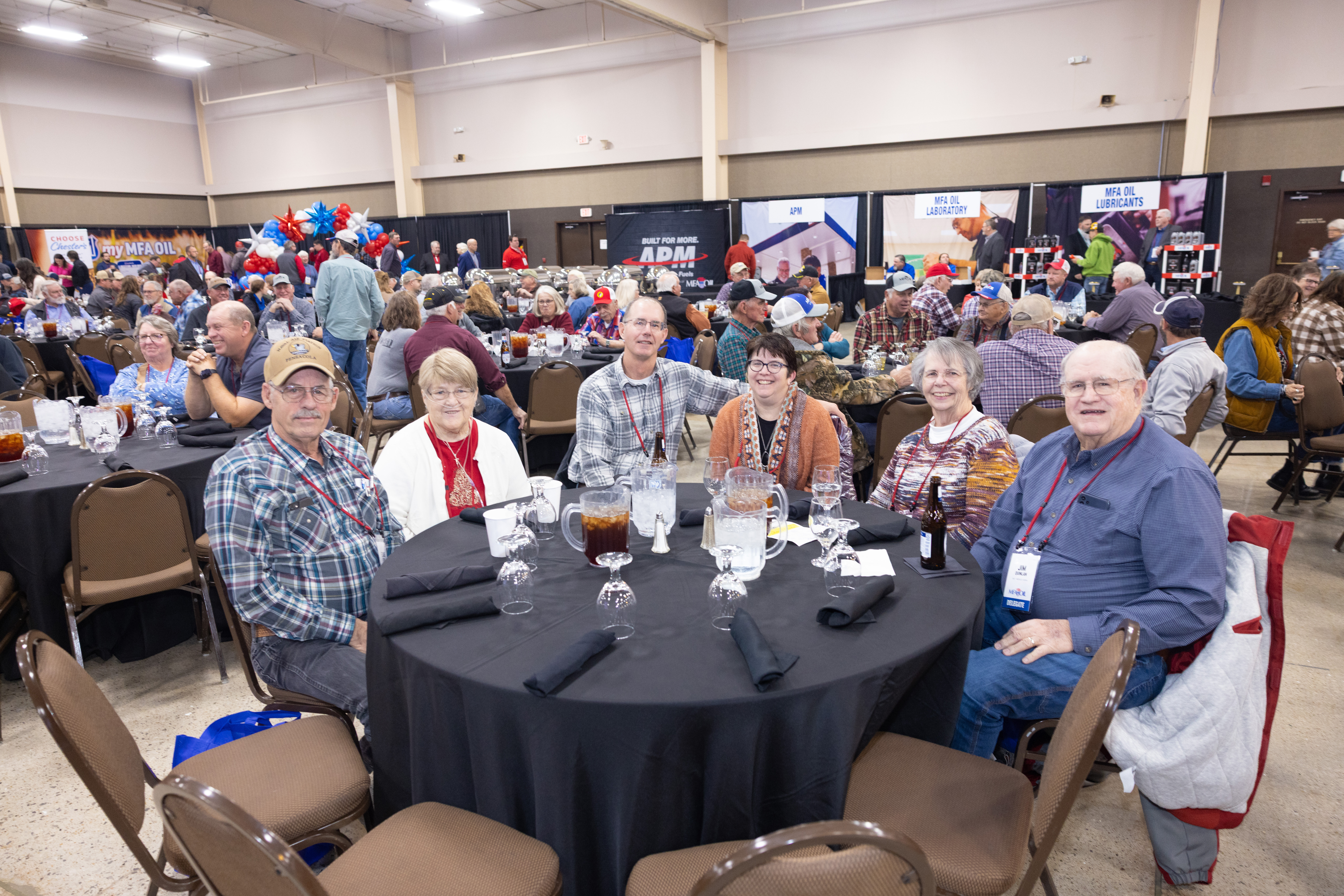 A group of seven people sits around a round table with black tablecloths at a large indoor event, with pitchers of drinks and many others seated at tables in the background.