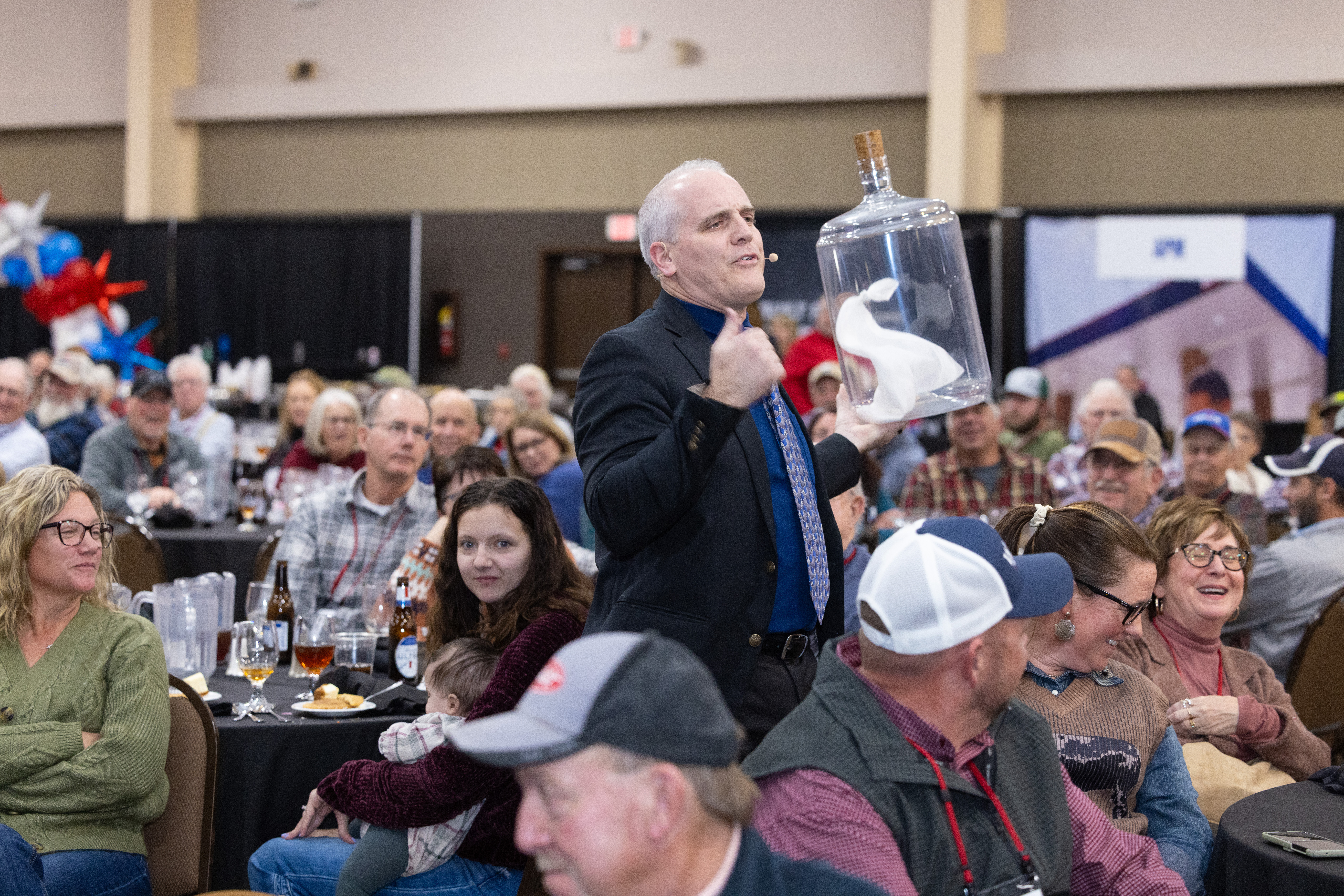 A man in a suit carries a large clear jar with a white cloth inside, walking among seated people at a crowded indoor event. The audience appears engaged and amused.