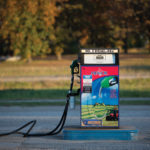 A lone diesel pump labeled No. 2 Diesel-Red stands on a concrete platform outdoors. The pump features a colorful advertisement and boasts a green nozzle with a long hose on the ground, against a backdrop of trees and grass.