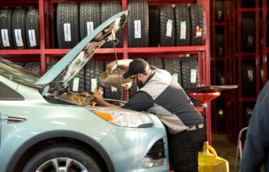 A mechanic pours oil into the engine of a light blue SUV with the hood open inside an auto shop, surrounded by shelves of stacked tires.