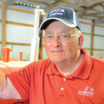 Elderly man wearing glasses, a cap, and a red polo shirt with a MAPCO OIL logo stands indoors next to farming equipment. Marion Kertz is seen against the backdrop of a metal-roofed structure.