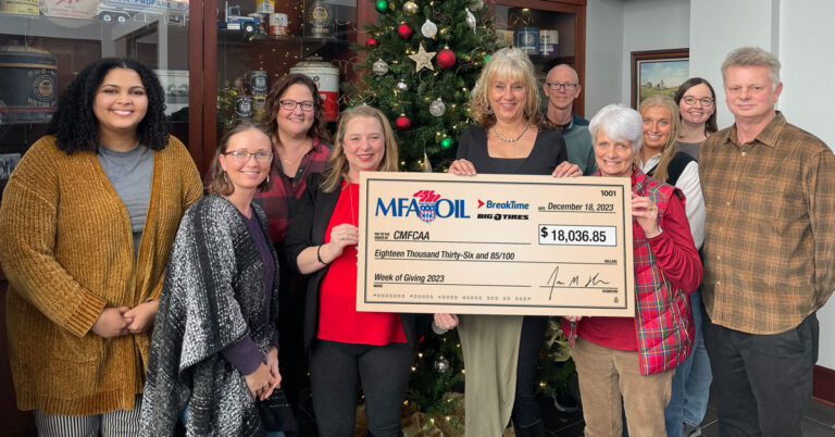 A group of people stand in front of a decorated Christmas tree, holding a large check for $18,036.85 from MFA Oil and Big Brothers Big Sisters, dated December 18, 2023. They are smiling and wearing casual attire.