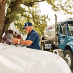 A person in a blue uniform and cap works on a large white propane tank outdoors, next to a blue truck. Theres a green tree and grassy area in the background.