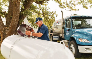 A person in a blue uniform and cap works on a large white propane tank outdoors, next to a blue truck. Theres a green tree and grassy area in the background.