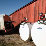 A red barn with two large white fuel tanks labeled MFA Oil and a parked fuel delivery truck partially visible, showing open compartments. The scene is lit by bright sunlight, casting clear shadows on the ground.
