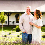 A smiling couple stands in a lush garden at Long Row Lavender, in front of a white, barn-like building. The man wears a light shirt and jeans while the woman, wearing a striped dress, gazes at him affectionately. Vibrant purple flowers bloom in the foreground.