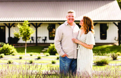 A smiling couple stands in a lush garden at Long Row Lavender, in front of a white, barn-like building. The man wears a light shirt and jeans while the woman, wearing a striped dress, gazes at him affectionately. Vibrant purple flowers bloom in the foreground.
