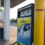 A gas station pump labeled MFA Oil offers unleaded fuel. The pump display shows a nozzle graphic under a clear sky. Surrounding the pump are other gas station equipment and a distant view of a fuel truck.