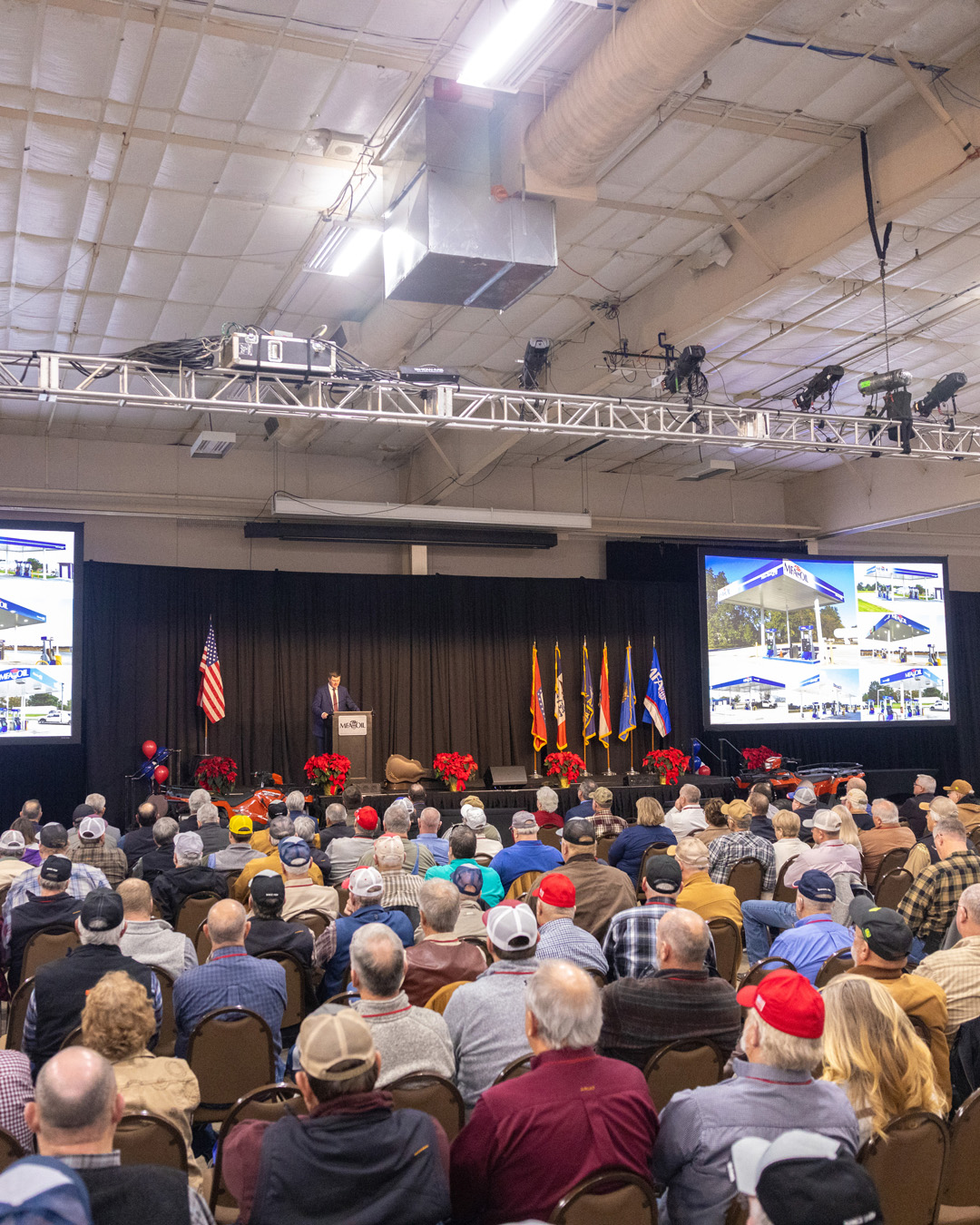 A large audience sits in rows facing a stage where a speaker stands at a podium. U.S. flags, military flags, and large screens displaying a building are behind the speaker in a spacious indoor venue.
