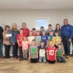 A group of children and adults standing indoors, with several children holding a certificate. One adult is holding a check. They are all smiling and casually dressed. The room has light-colored walls and hardwood floors.