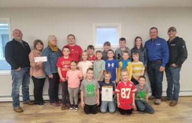 A group of children and adults standing indoors, with several children holding a certificate. One adult is holding a check. They are all smiling and casually dressed. The room has light-colored walls and hardwood floors.